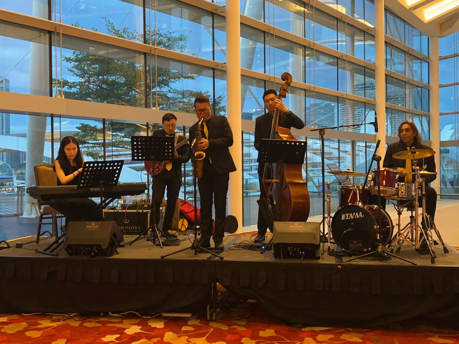 Group of people playing musical instruments on a stage in front of a large glass wall, Live Bands