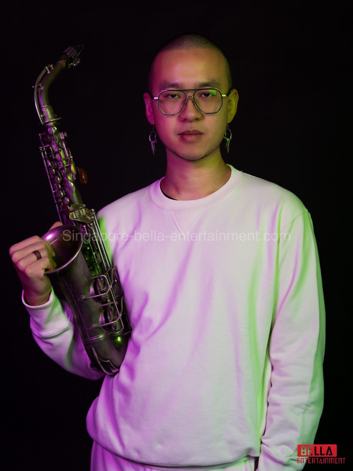 Man wearing a white t-shirt and spectacles, holding a saxophone in his hand against a black background, Saxophonist