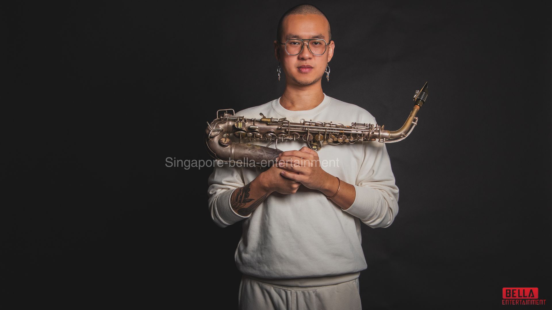 Man wearing a white t-shirt and spectacles, holding a saxophone in his hands against a black background, Saxophonist