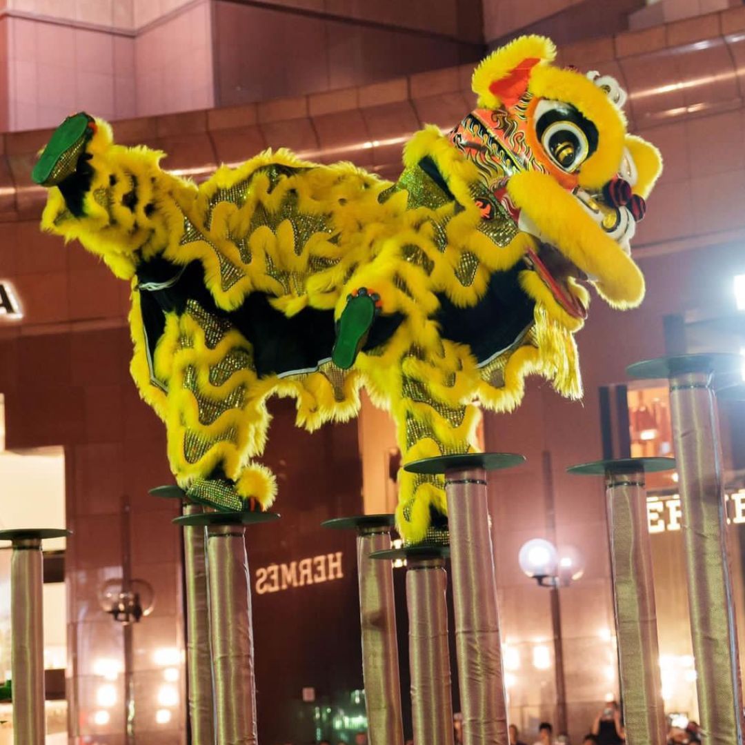 Yellow lion statue in front of a building, surrounded by people, poles, lights, boards with text, and other, Chinese Entertainment Singapore for Hire
