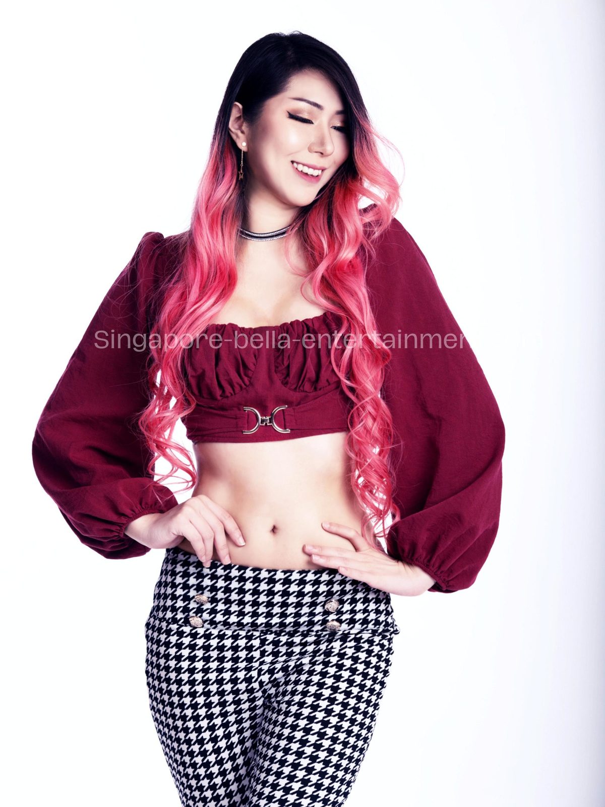 Woman with pink hair wearing a maroon top and black and white houndskin pants, standing and smiling against a, Hire DJ Singapore