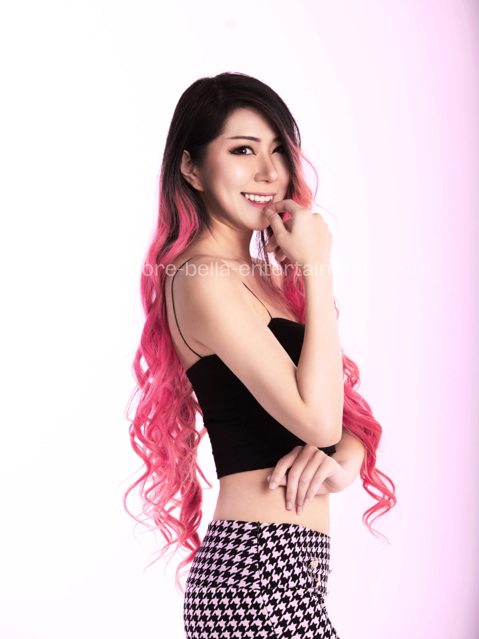 Woman with long pink hair wearing a black top and houndskin skirt, standing and smiling against a white, Hire DJ Singapore