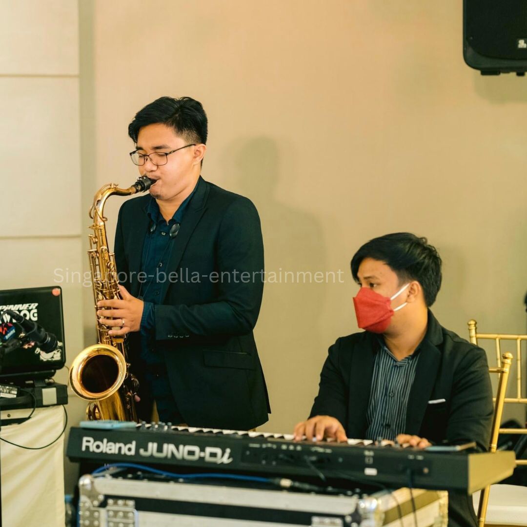 Man playing a saxophone in front of a keyboard, with another person sitting on a chair to his right, Saxophonist