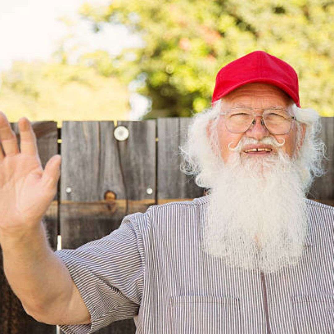 Older man with a long white beard and a red hat, wearing spectacles and smiling, standing in front of a, Impersonator for Hire