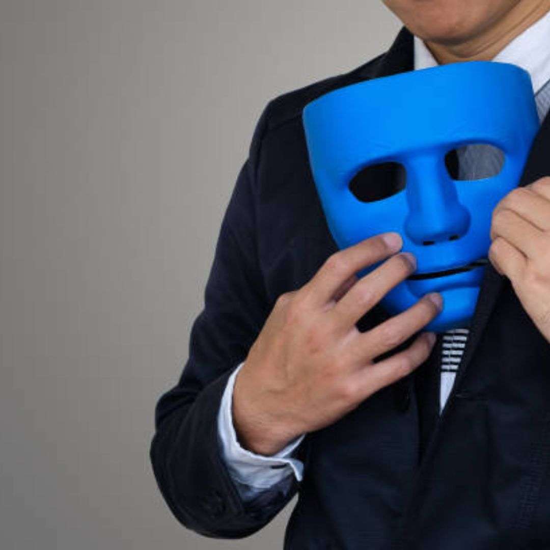 Man in a suit holding a blue mask in front of his face, with a wall in the background, Impersonator for Hire