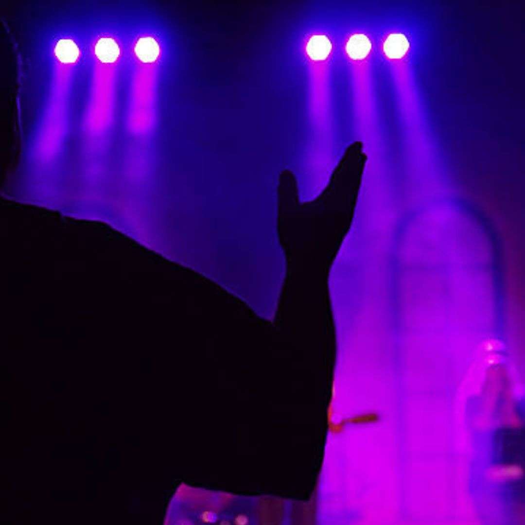 Man standing in front of a stage with his hands up in the air, illuminated by a few lights at the top of the, Musicians