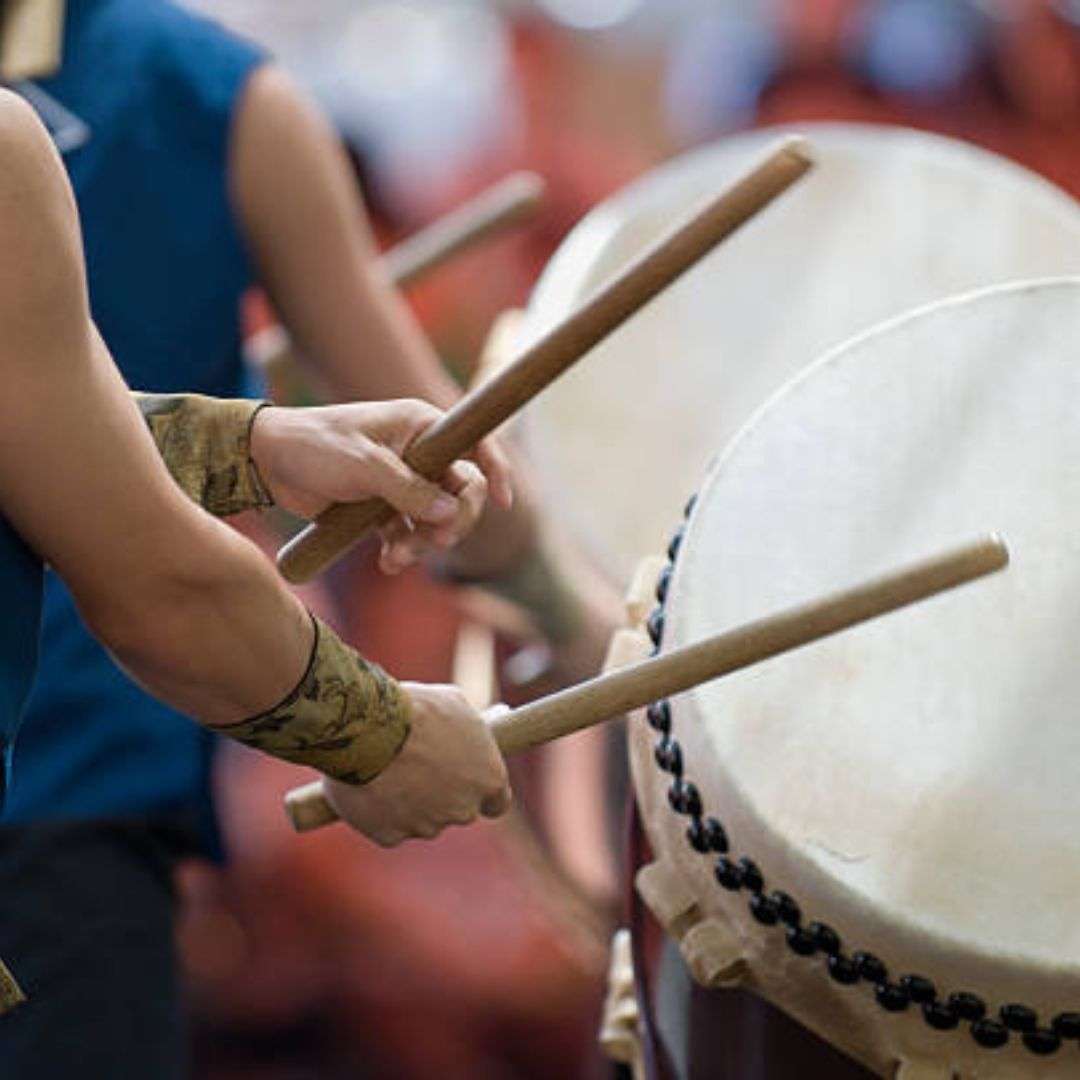 Group of people playing drums with sticks in their hands, likely part of a drumming class at a festival, Musicians