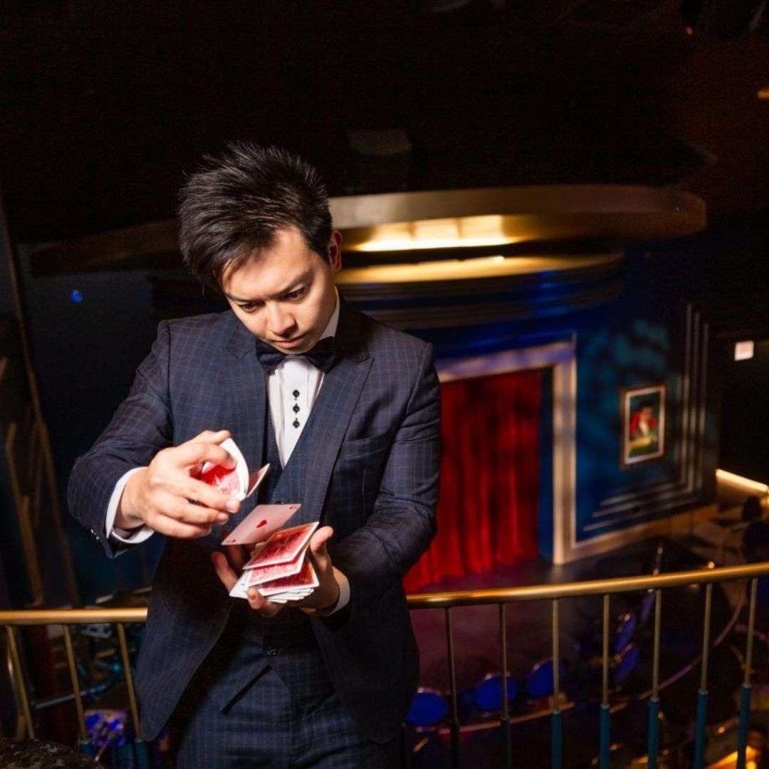 Man in a tuxedo holding a deck of cards, standing in front of a railing with a few people in the background, Hire Birthday Entertainment in Singapore