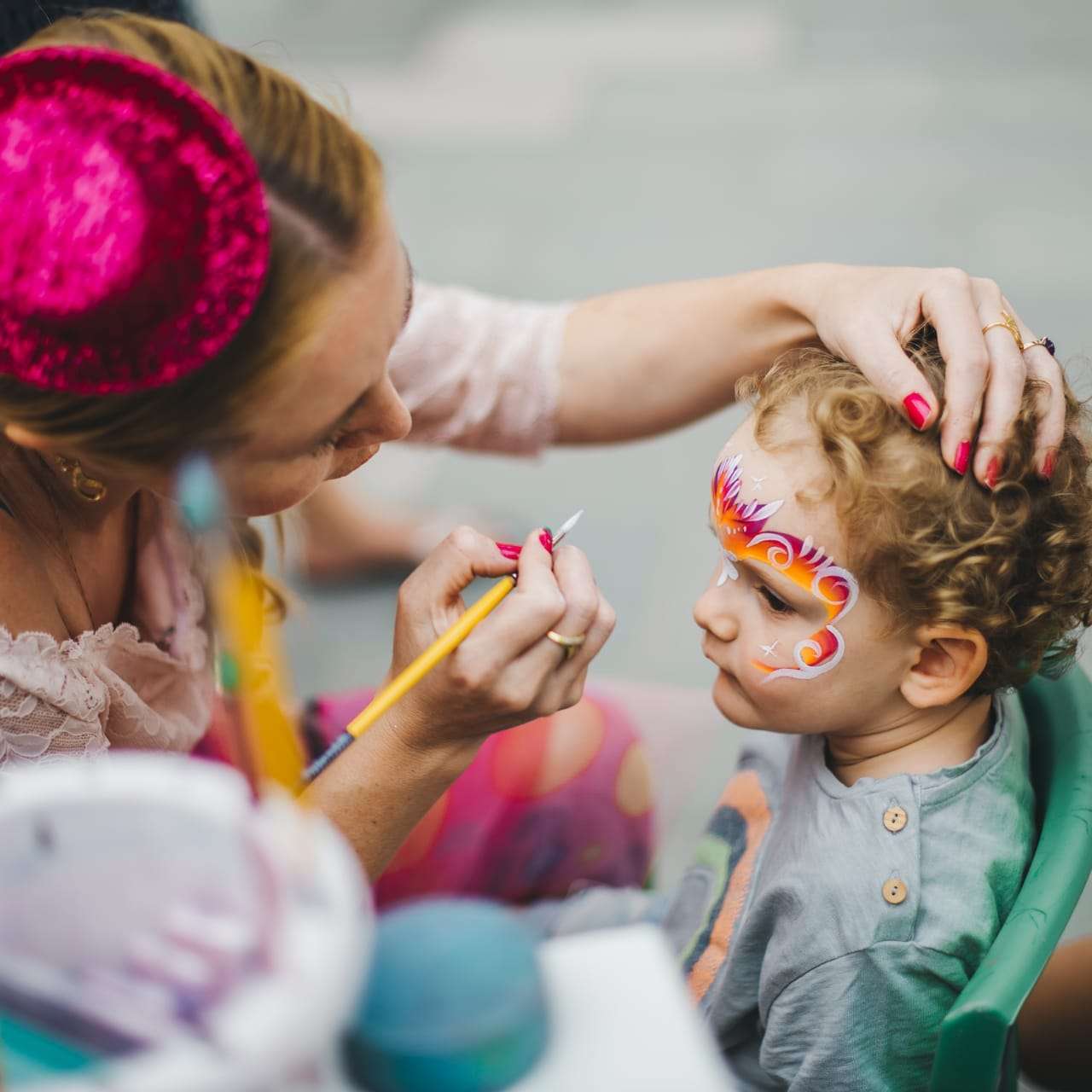 Little girl sitting in a chair with her face painted, while a woman stands beside her holding a brush and, Hire Birthday Entertainment in Singapore