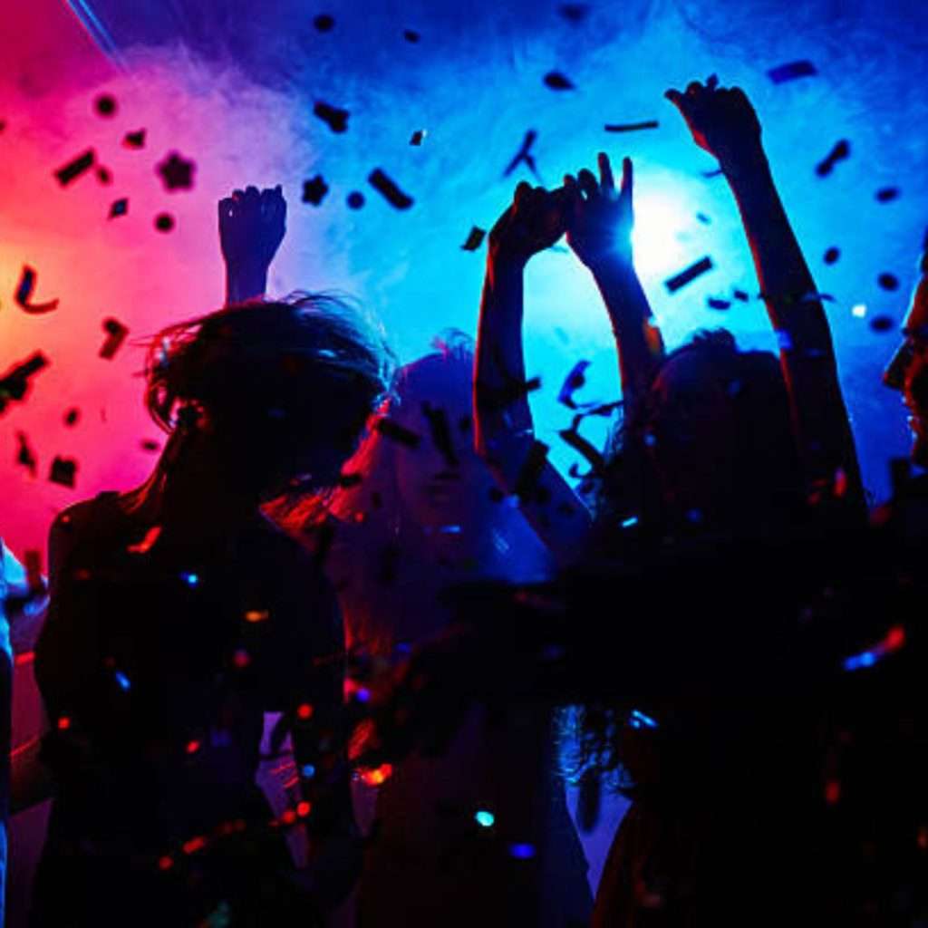 Group of people dancing at a party with confetti in the air, illuminated by colorful lights in the background